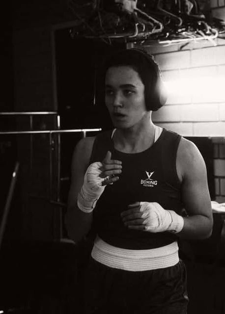 Person in boxing gear standing in a gym with boxing equipment in the background.